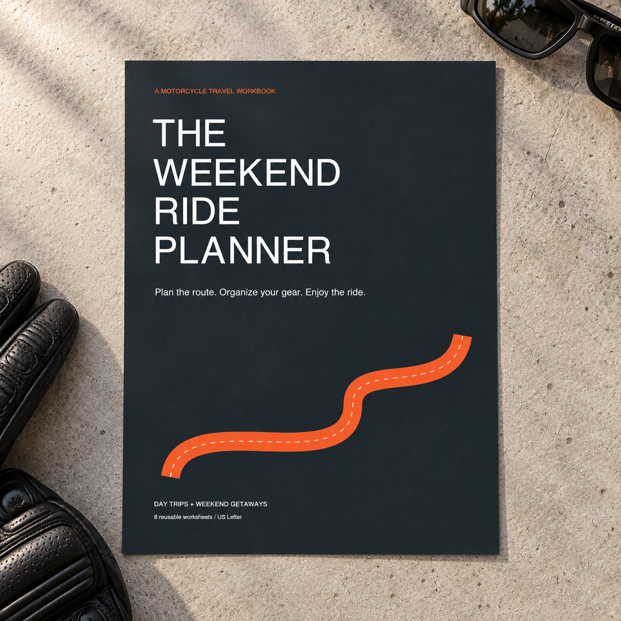 Weekend Ride Planner printable cover on a concrete surface, styled with riding gloves and glasses. Props not included.